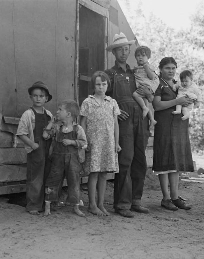 A family from history stands barefoot outside a wooden structure, depicting life during historical events.