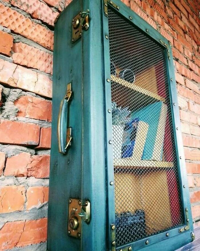 Repurposed suitcase shelf with mesh door, mounted on a brick wall, showcasing creative recycling.