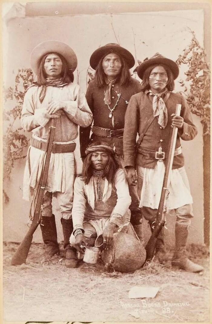 Historical image of four Apache men, three standing with rifles and one sitting, showcasing cultural attire.