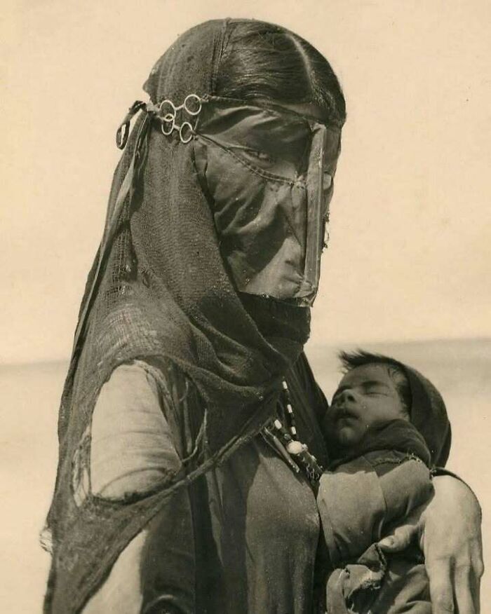 Woman in historical attire with intricate face covering holding a sleeping baby, showcasing something interesting in history.