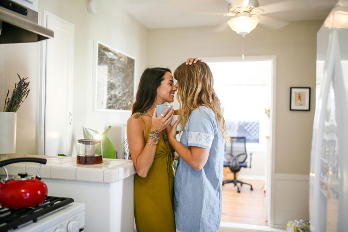 Two women standing closely in a kitchen, one holding a mug, highlighting support in a wedding dilemma for a gay woman. Two women standing closely in a kitchen, one holding a mug, highlighting support in a wedding dilemma for a gay woman.