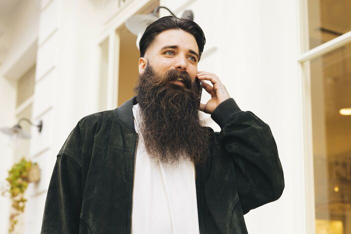Man with a long beard talking on the phone, wearing a black jacket and white shirt, illustrating recent fashion trends people hate.