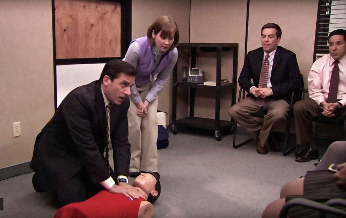 Scene of a man comically attempting CPR on a dummy, with onlookers in an office, highlighting unrealistic movie moments.