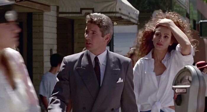 A couple walking outdoors in 90s fashion, with the woman adjusting her hair and the man wearing a suit.