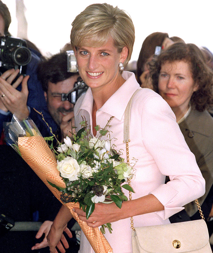 "Smiling celebrity in a pink suit holding flowers, surrounded by people and photographers."