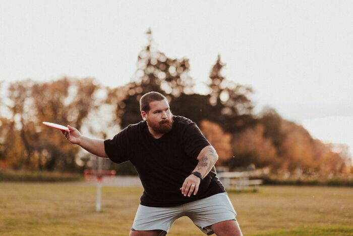 A man playing frisbee outdoors, highlighting unconventional beauty against a backdrop of trees and a clear sky.