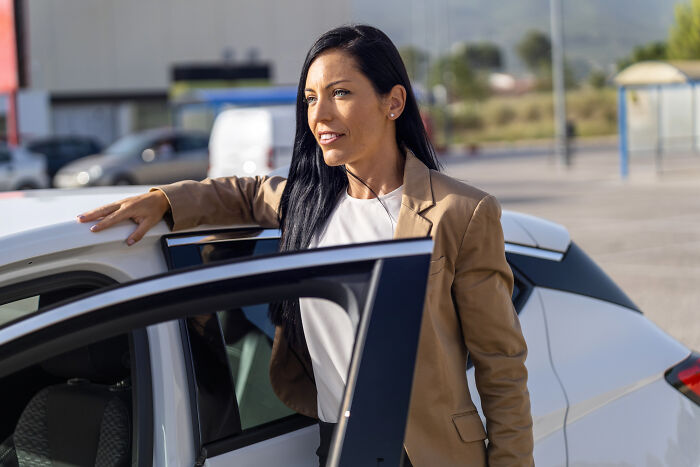 Woman in a brown blazer stands beside a car, challenging job assumptions with a confident smile.