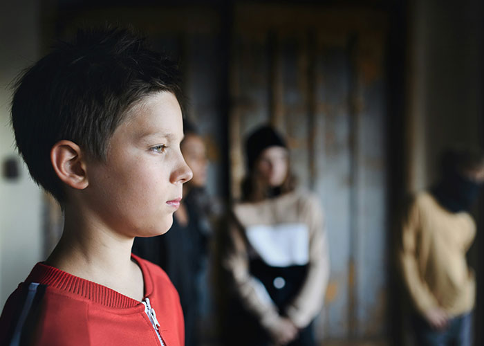Young boy looking serious, wearing a red shirt, with blurred stepsiblings in the background during a family gathering.