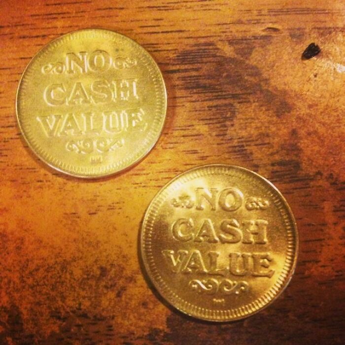 Two tokens on a wooden surface with "No Cash Value" engraved, reflecting mildly infuriating server tips.