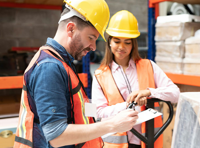 Two workers in hard hats and vests discussing a checklist in a warehouse.