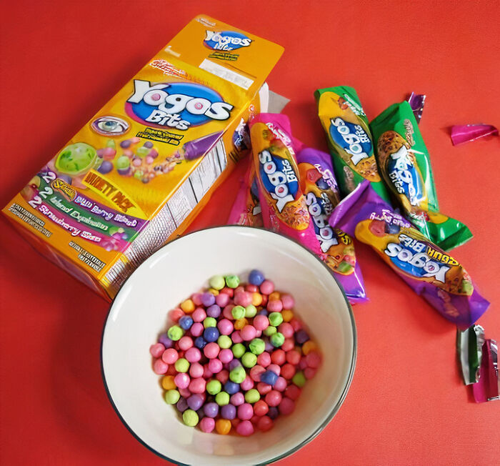 Colorful Yogos Bits from 90s and 2000s in bowl, next to packaging and wrappers on red background.