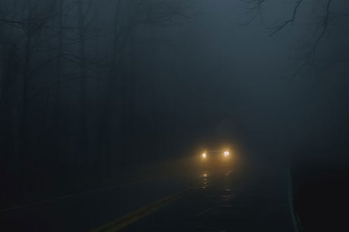 Car driving with headlights on through a foggy night, possibly highlighting people doing something wrong.