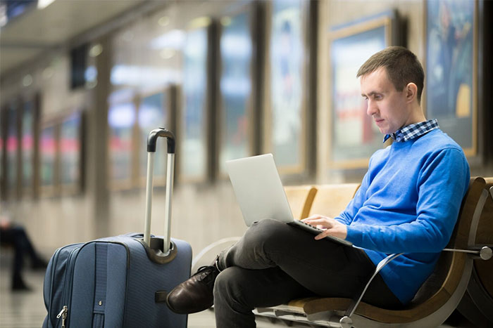Man in a blue sweater using laptop on bench at train station with suitcase, realizing he might be doing something the wrong way.