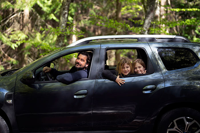 Man driving with two children smiling in the backseat, surrounded by forest scenery, representing family and adventure.