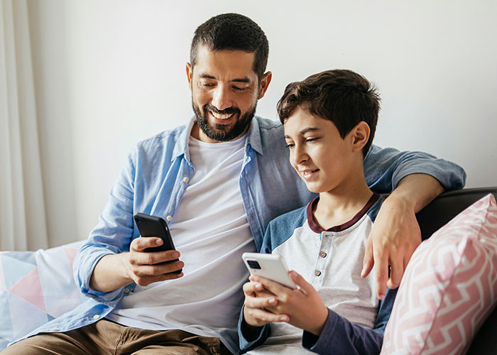 Man with his child sitting on a couch, both smiling while using their smartphones, highlighting bonding moments. Man with his child sitting on a couch, both smiling while using their smartphones, highlighting bonding moments.