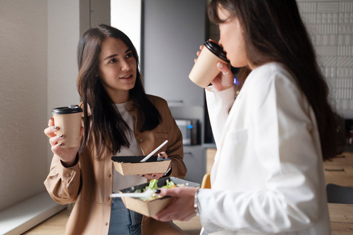 Two coworkers having a discussion at work, one holding a coffee cup and a food container, highlighting team spirit.