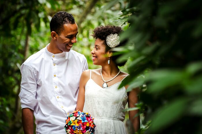 Happy couple in wedding attire smiling at each other, surrounded by greenery.