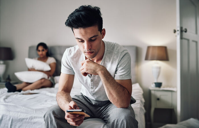 Man sitting on bed looking at phone, appearing thoughtful; woman in background. Concept of regret in marriage and spouse relationships.