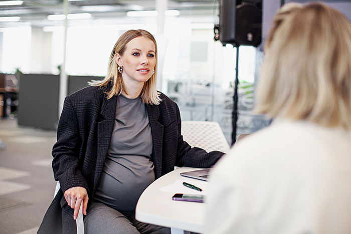 Pregnant coworker in a gray dress and blazer sitting at a table, talking to a colleague in an office setting. Pregnant coworker in a gray dress and blazer sitting at a table, talking to a colleague in an office setting.