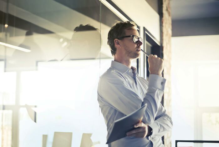 Person in glasses pondering at a window, holding paperwork in an office setting, embodying traits often seen in discussions about rich people.