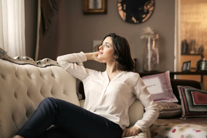 A woman in a white blouse sitting on a luxurious sofa, gazing thoughtfully, capturing a sense of rich ambiance.