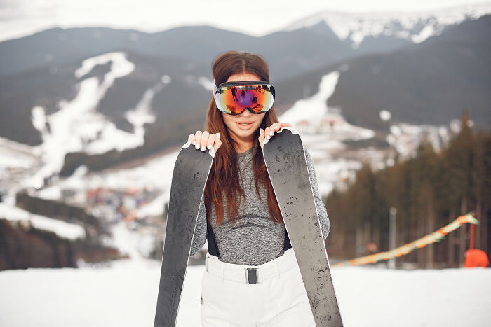 Woman holding skis on a snowy mountain, wearing goggles, embodying a stereotype of rich people leisure.