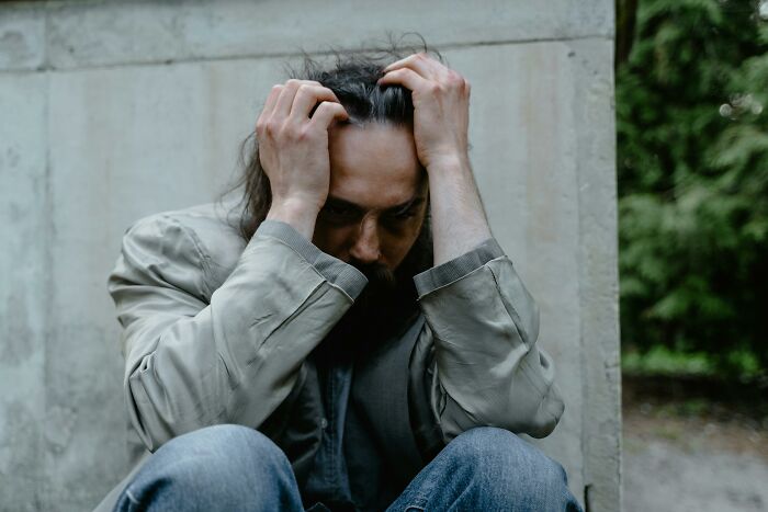 Person sitting with head in hands, appearing frustrated or upset at rich people, wearing a gray jacket and jeans. 