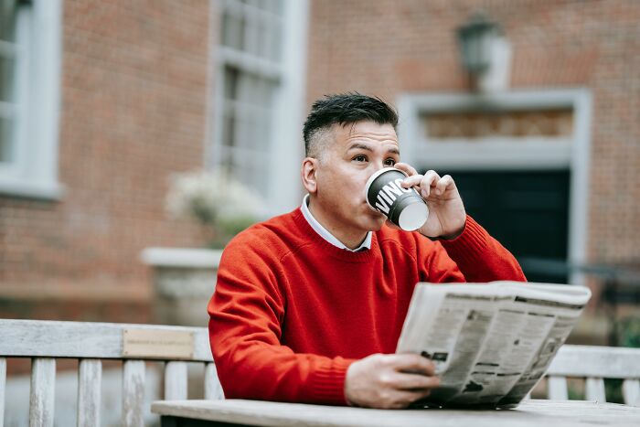 Person in a red sweater sipping coffee and reading a newspaper, depicting rich people's annoying habits.