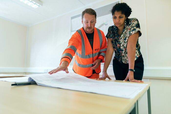 Two people examining blueprints at a table, with one wearing a reflective jacket, discussing project details indoors.