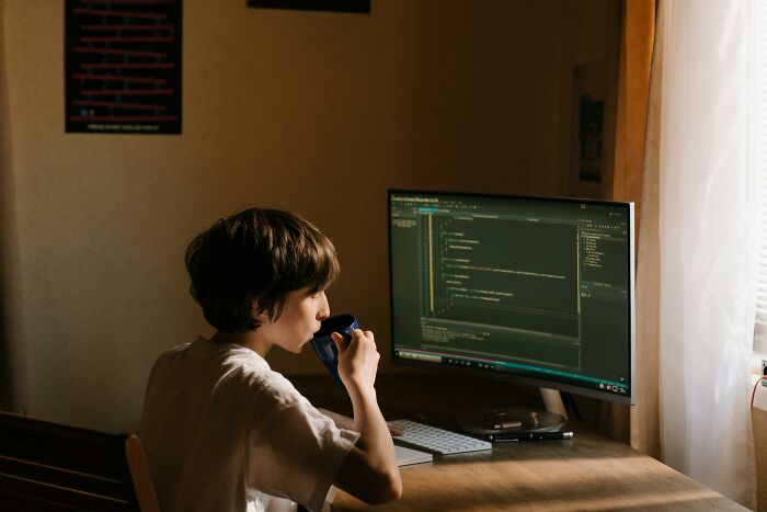 A child drinking from a mug, sitting at a desk with a computer screen displaying coding, symbolizing stories of overnight wealth.