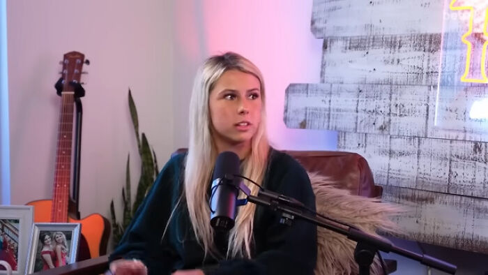 A woman speaking into a microphone during a podcast on absurd stories of overnight wealth, with guitar in the background.