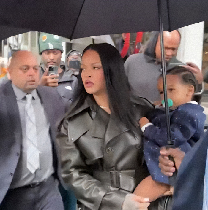 Rihanna holding her child, under an umbrella, surrounded by people in a public setting. Rihanna holding her child, under an umbrella, surrounded by people in a public setting.