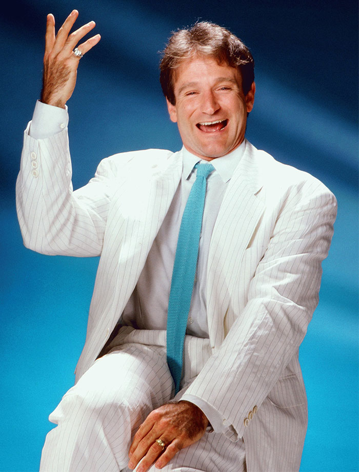 Celebrity in a white suit with a blue tie, smiling and gesturing against a blue background.