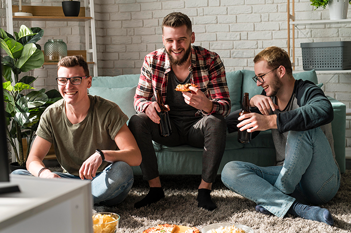 Three roommates laughing, sitting on the floor with snacks and drinks, enjoying a casual gathering.