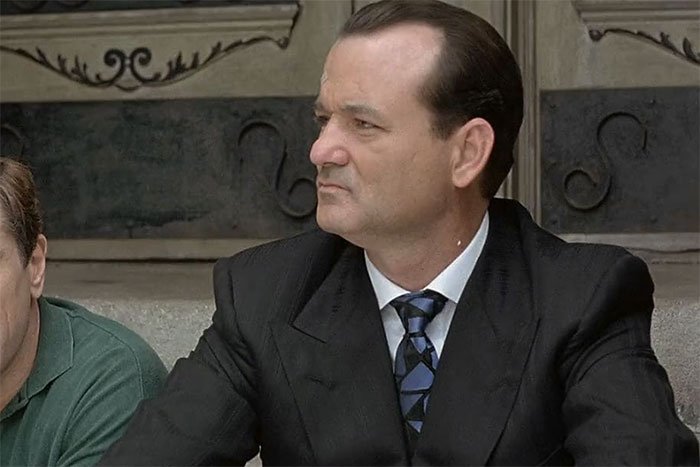 A person in a suit looks away with a serious expression, sitting outdoors.