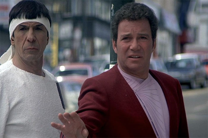 Two actors in a city street scene, one wearing a headband, the other in a red jacket, from a sci-fi film.