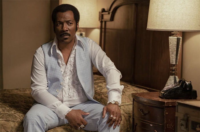 Actor in vintage attire sitting on a bed, illuminated by two lamps on side tables.