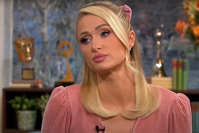 Celebrity in a pink dress sits in a studio with awards visible in the background.