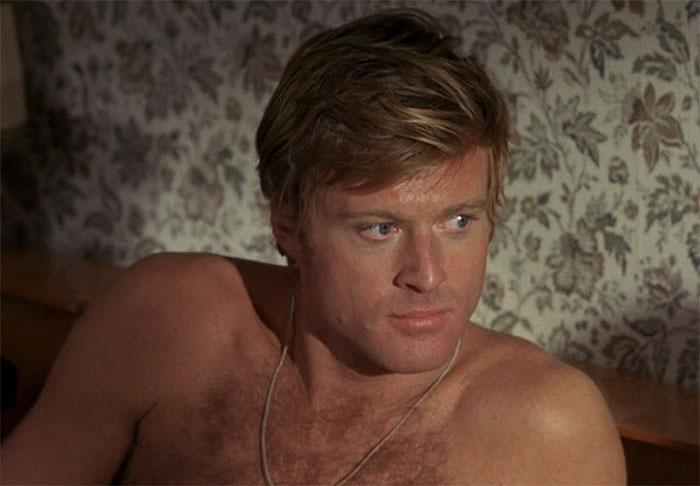 A shirtless actor with blonde hair looks intently off-camera, set against a floral wallpaper backdrop.