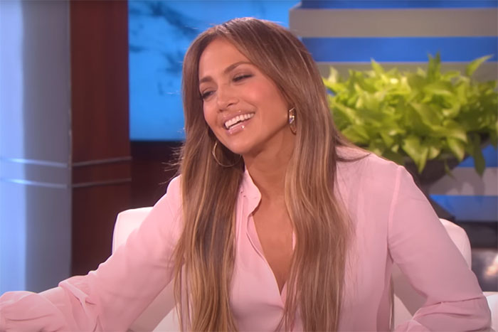 Celebrity smiling in a talk show, wearing a pink blouse, seated against a backdrop with plants.