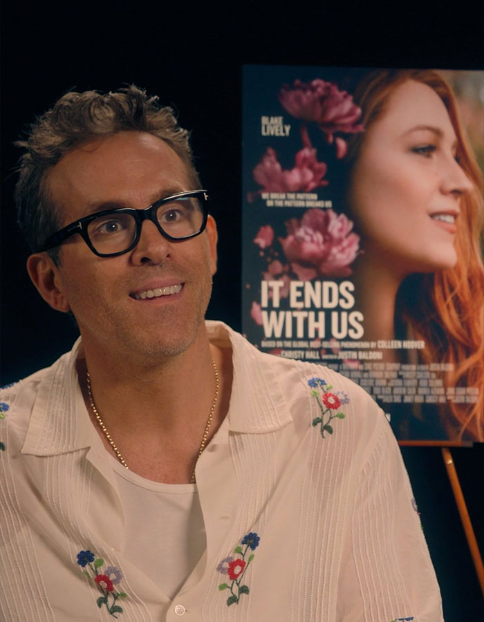 Actor at a press event wearing glasses and floral shirt, with movie poster in the background. Actor at a press event wearing glasses and floral shirt, with movie poster in the background.