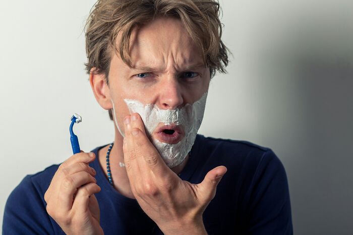 Man with shaving cream on his face, holding a razor, looking surprised, possibly thinking about unique gift ideas.