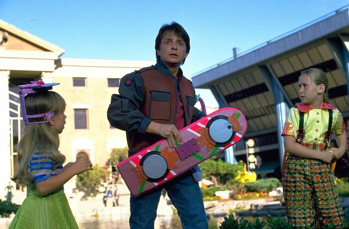 Man holding a famous movie prop hoverboard, standing between two children outside futuristic buildings.