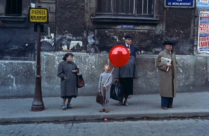 A child holding a red balloon stands on a sidewalk, an iconic famous movie prop in a vintage street scene.