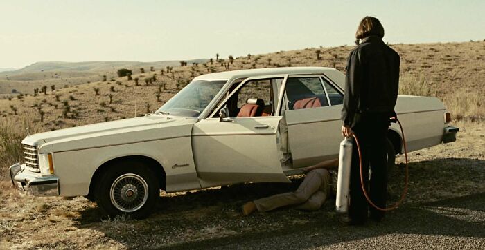 A man with a tank stands by a vintage car in a desert setting, depicting a famous movie prop scene.
