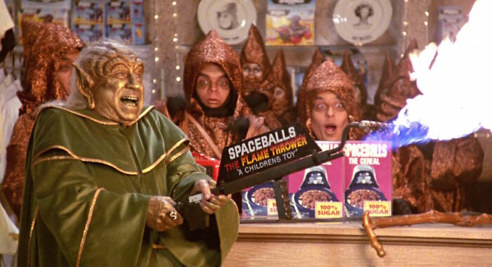 Spaceballs character using a humorous flame thrower, surrounded by cereal boxes in a scene from the movie.