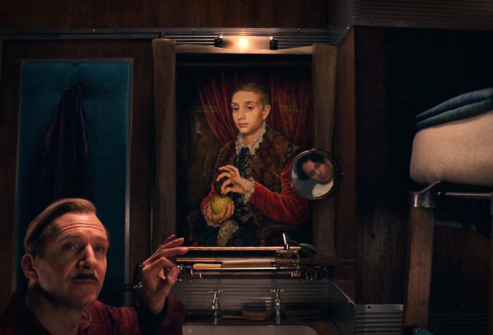 A man in front of a painting, holding a brush, inside a vintage train cabin.