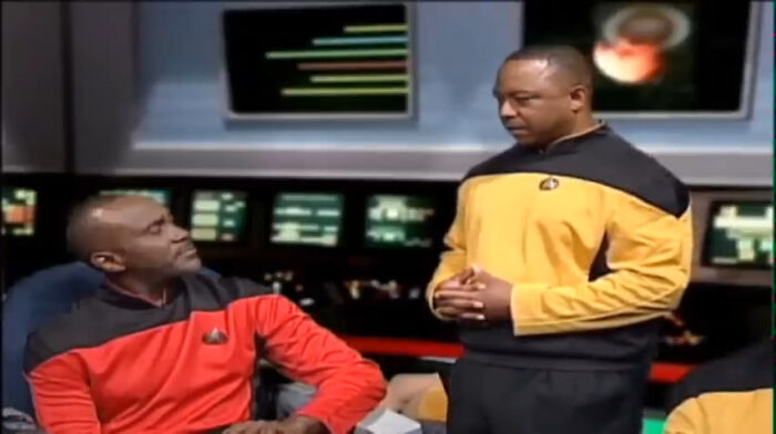 Two men in futuristic uniforms on a bridge set, possibly discussing weird government projects.