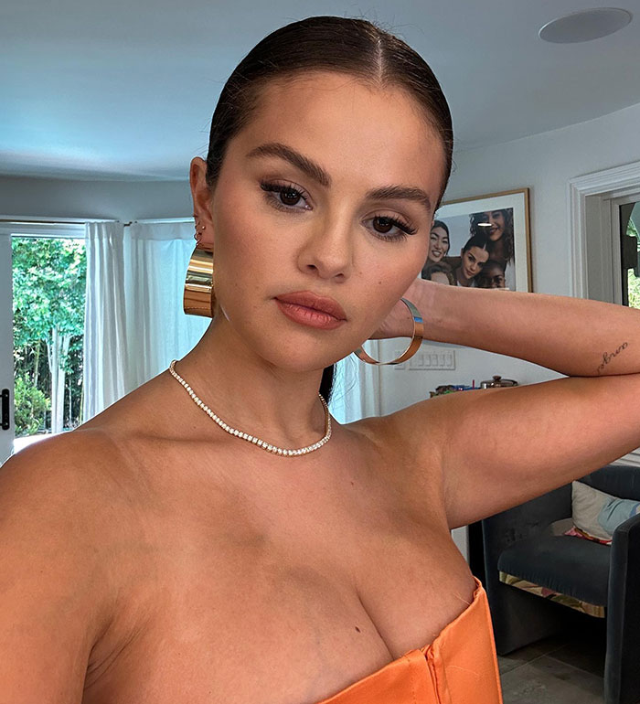 Selena Gomez showcasing weight loss, wearing a strapless orange dress and gold hoop earrings in a living room setting.