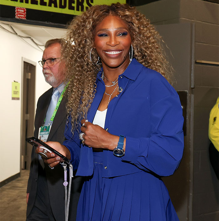 Woman in blue outfit smiles backstage with phone, curly hair styled, near others in a hallway. Woman in blue outfit smiles backstage with phone, curly hair styled, near others in a hallway.